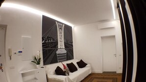 Una habitación con un sofá blanco, un escritorio pequeño, un ventanal con vistas a un puente y una alfombra estampada.