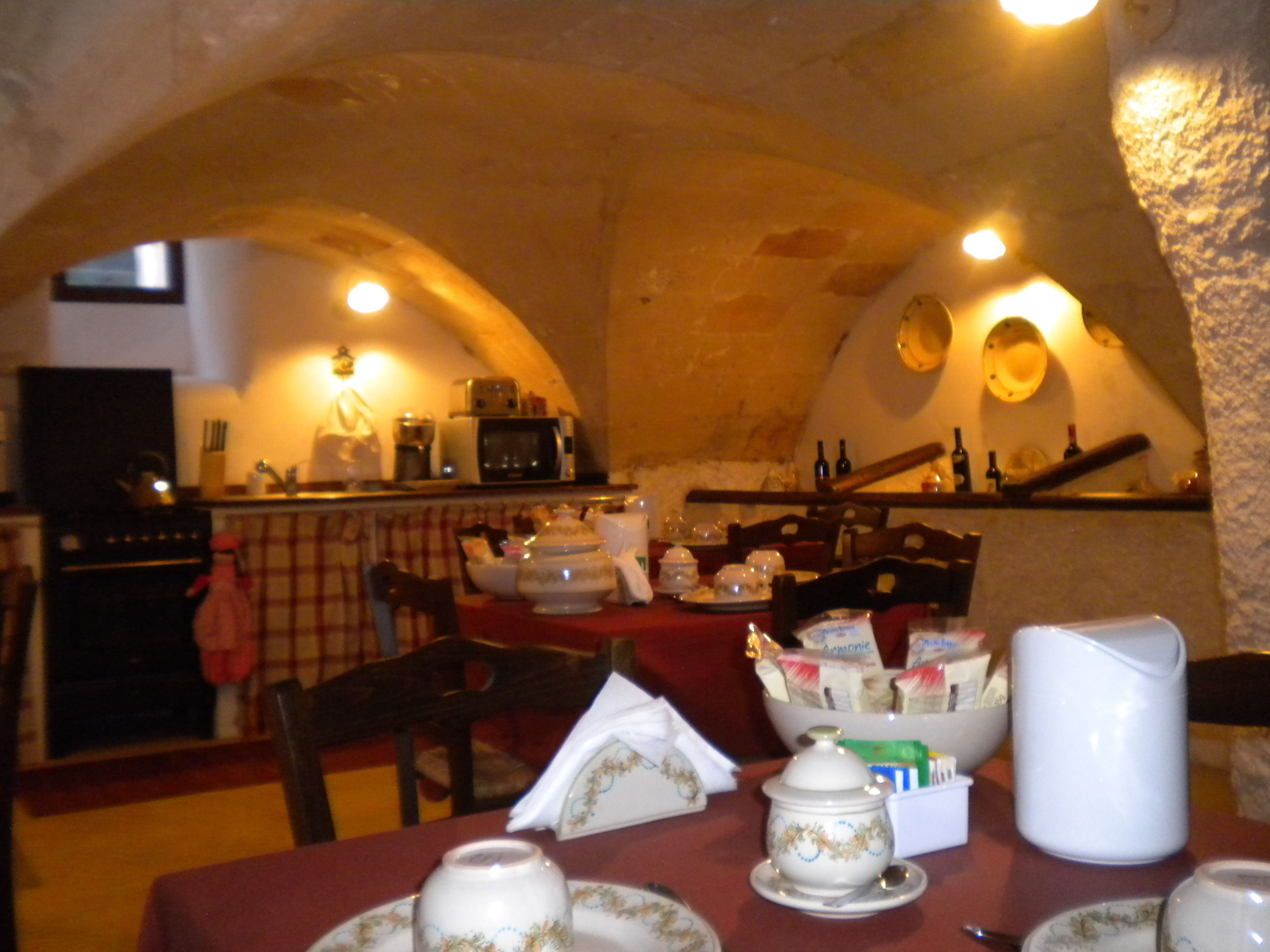 A dining area with a table set for four, featuring a variety of dishes and a pitcher. The room has a rustic stone ceiling and a kitchen in the background.