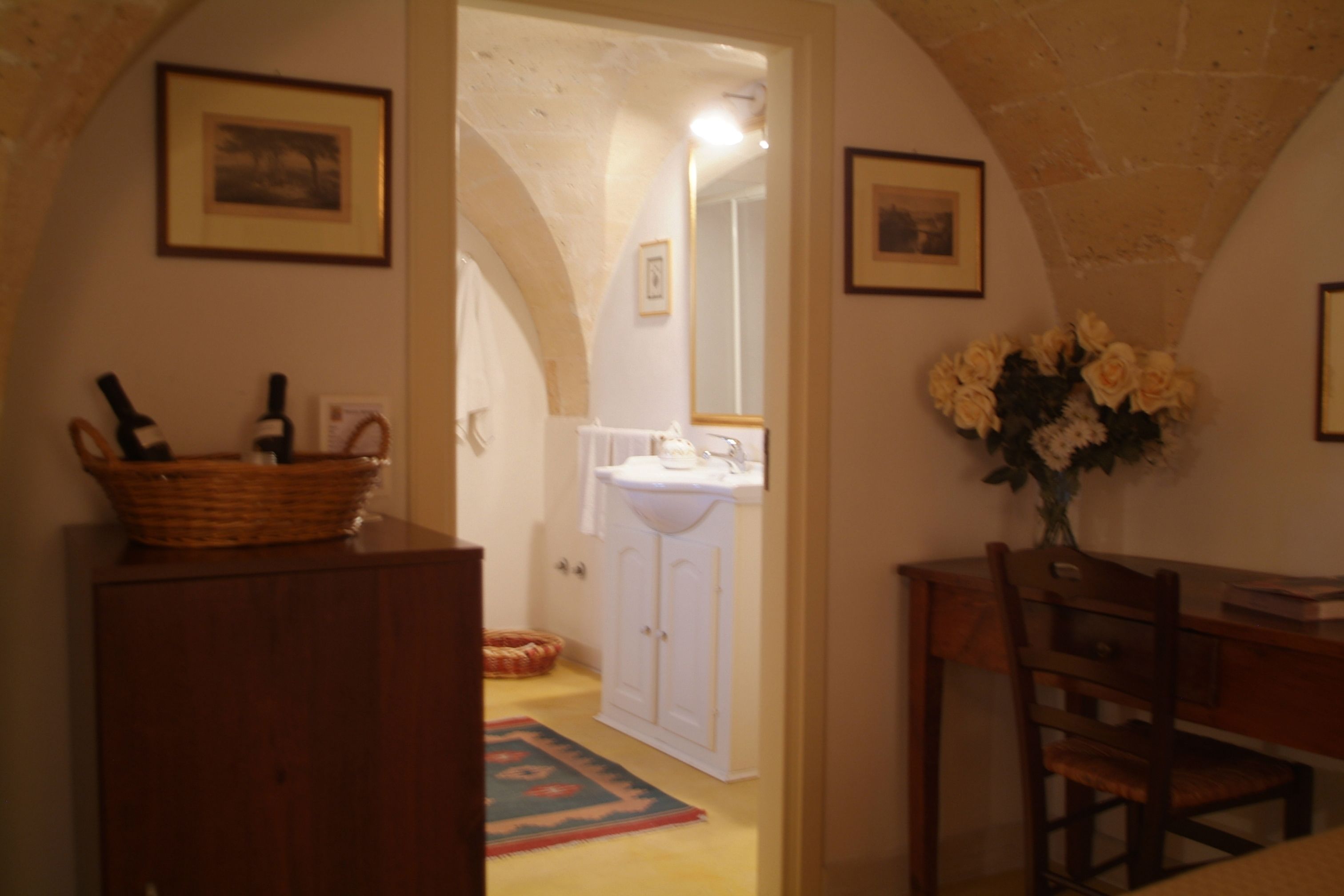 Un intérieur comprenant un bureau en bois, une chaise, un panier contenant des bouteilles, un vase avec des fleurs et une porte donnant sur une salle de bain équipée d’un lavabo et d’un miroir.