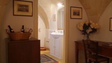 Un intérieur comprenant un bureau en bois, une chaise, un panier contenant des bouteilles, un vase avec des fleurs et une porte donnant sur une salle de bain équipée d’un lavabo et d’un miroir.