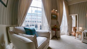A well-lit room with a white sofa, a small table with a lamp, and a view of buildings through large windows.
