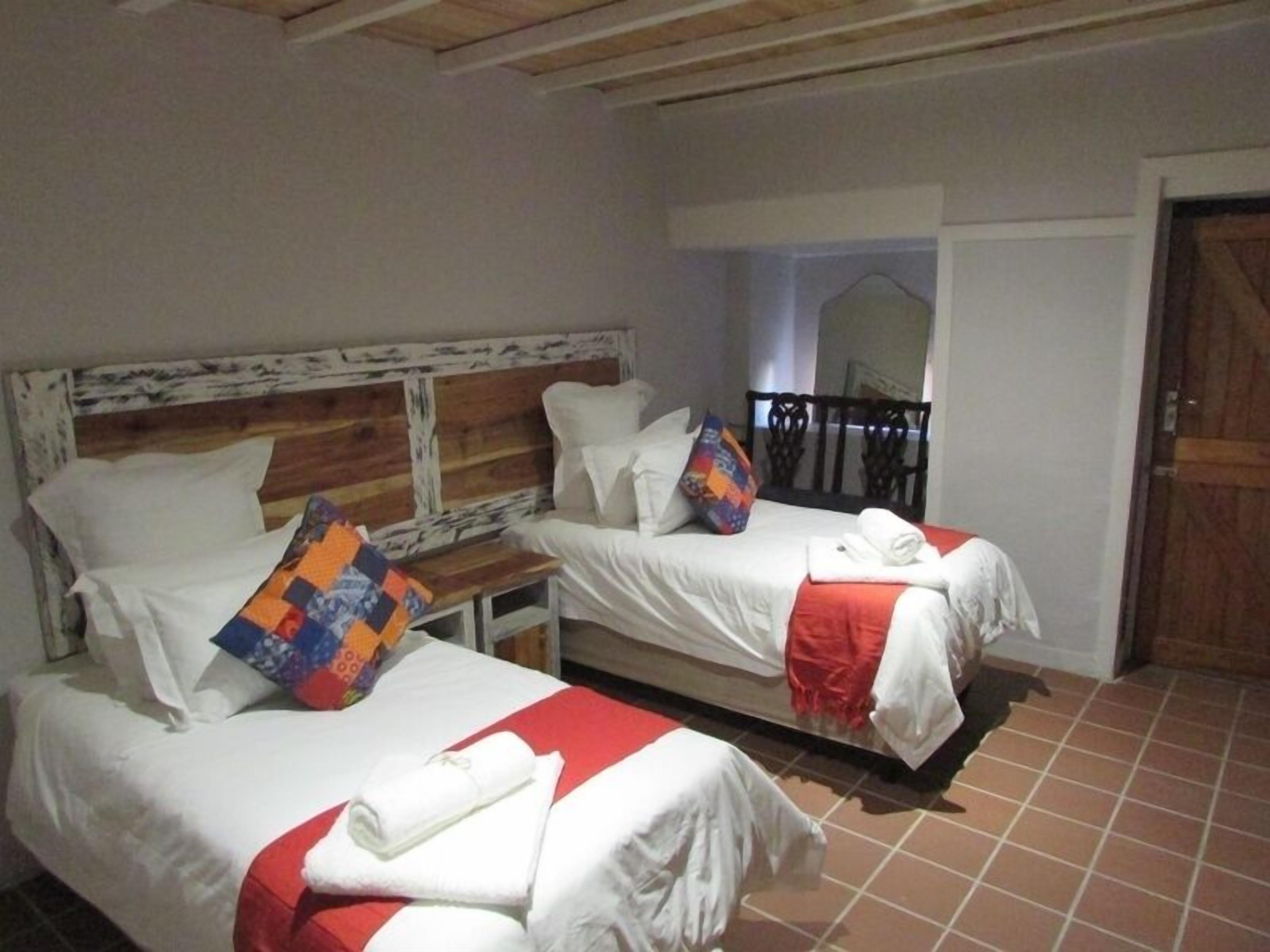 Une chambre d’hôtel avec deux lits, chacun agrémenté de draps blancs et d’un élément décoratif rouge. Un lit est doté d’une tête de lit en bois, l’autre d’une tête de lit en métal. Un couvre-lit coloré recouvre le lit muni de la tête de lit en bois.