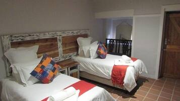 Une chambre d’hôtel avec deux lits, chacun agrémenté de draps blancs et d’un élément décoratif rouge. Un lit est doté d’une tête de lit en bois, l’autre d’une tête de lit en métal. Un couvre-lit coloré recouvre le lit muni de la tête de lit en bois.