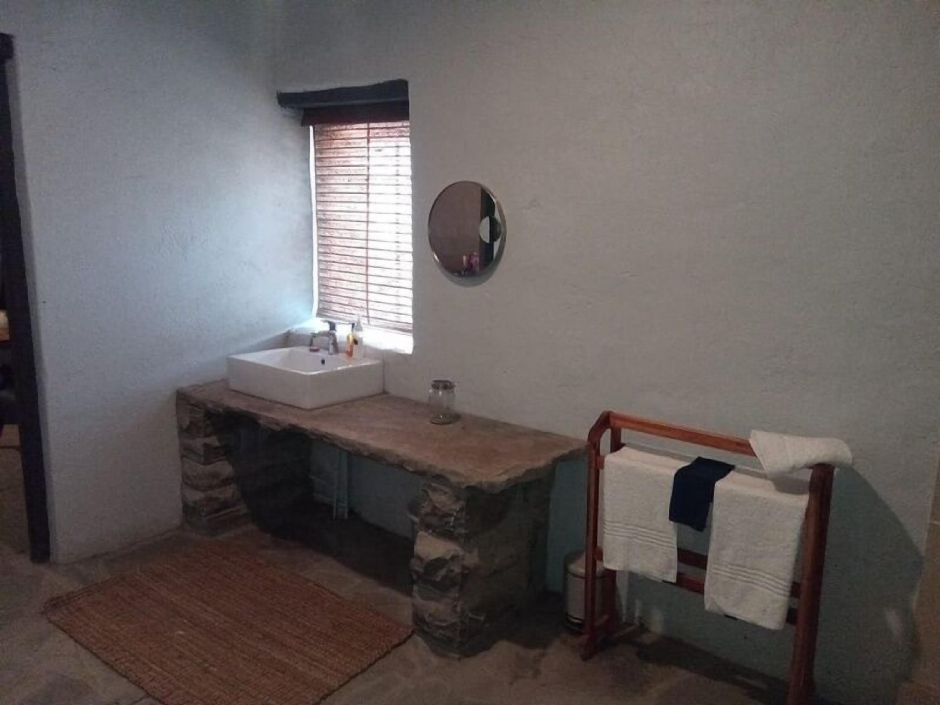 A bathroom with a stone sink, a mirror, a window with blinds, a wooden bench with towels, and a rug on the floor.