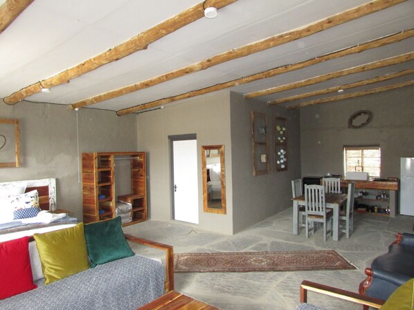 A rustic interior with exposed wooden beams, a stone floor, a sofa with colorful cushions, a dining area with chairs, and a kitchen area with a refrigerator. - Rogge Cloof (Karoo Hoogland)