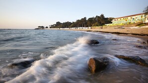 Un hotel frente a la playa con una hilera de habitaciones a lo largo de la costa, una playa de arena y olas rompiendo contra las rocas.