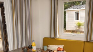 Un área de comedor con una mesa dispuesta para dos personas, un sofá amarillo y vistas a una casa a través de una ventana.