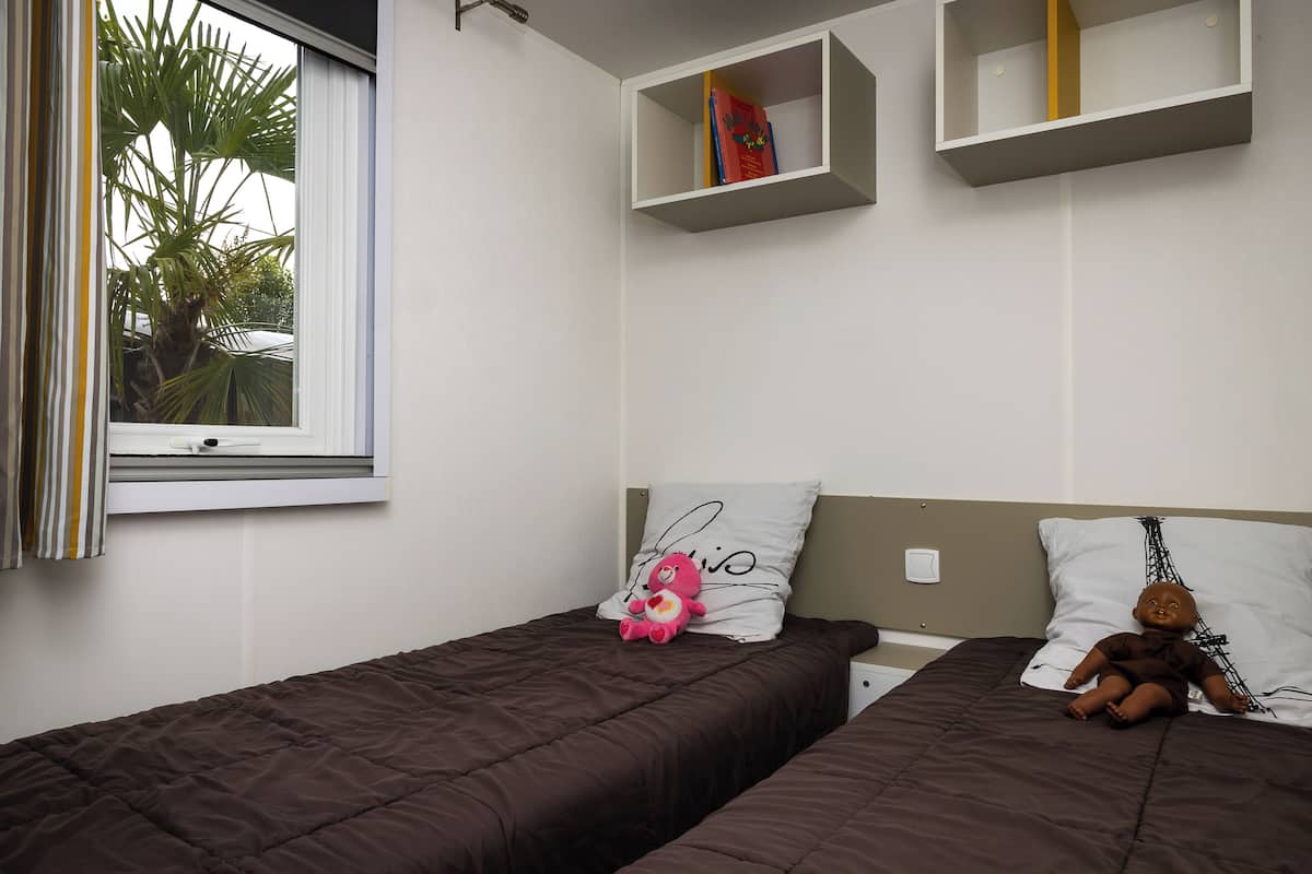 A bedroom with two beds, a window with curtains, and a shelf with books.