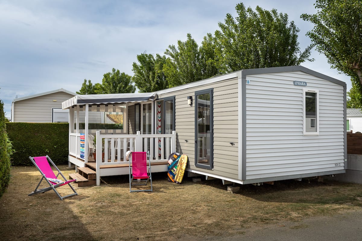 A mobile home with a covered porch, a deck, and a small garden area with a lawn chair and a surfboard.