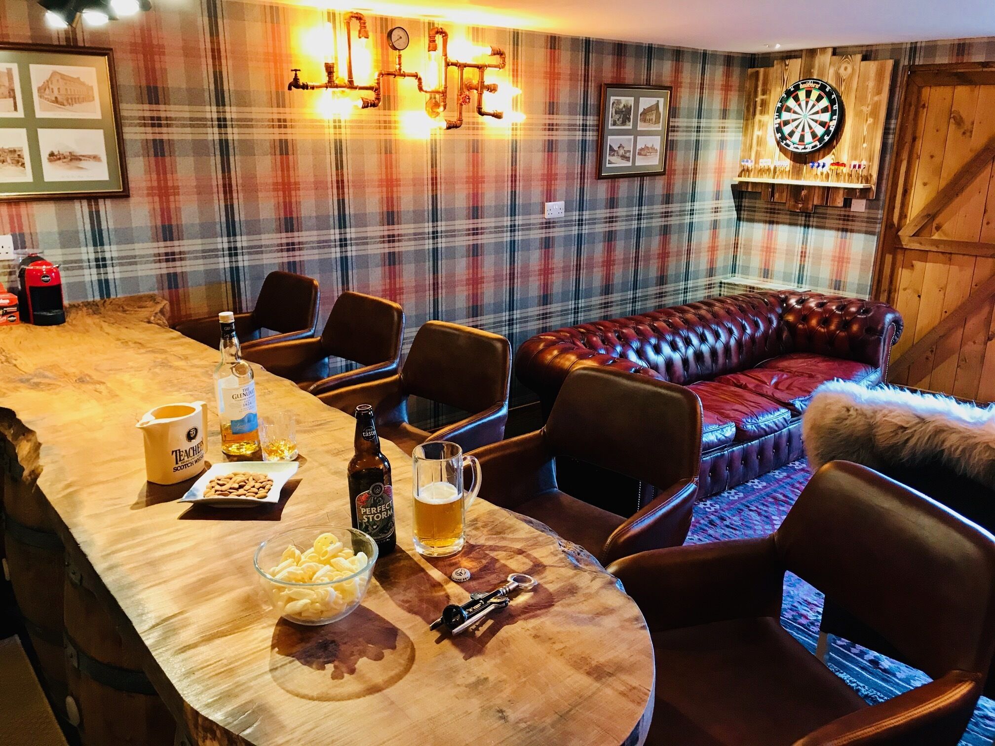 Une pièce chaleureuse avec un papier peint à carreaux, un canapé en cuir, une table en bois sur laquelle sont disposés des boissons et des en-cas, et un jeu de fléchettes accroché au mur.