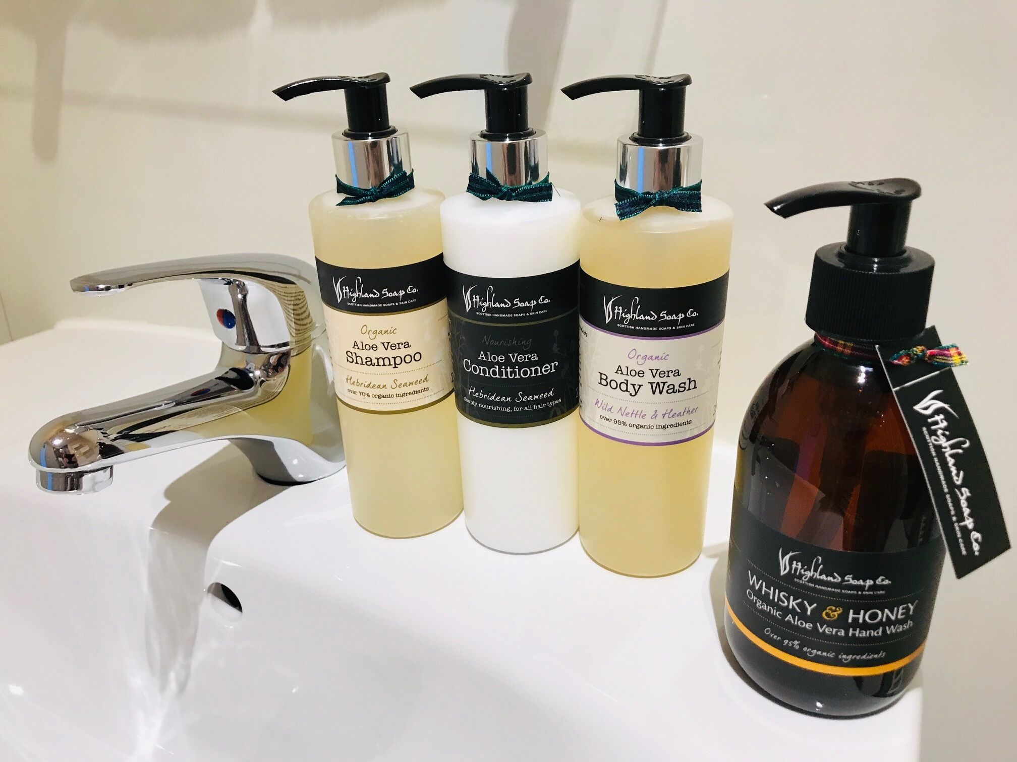A bathroom sink with four bottles of Highland Soap Co. products: Organic Aloe Vera Shampoo, Nourishing Aloe Vera Conditioner, Organic Aloe Vera Body Wash, and Whiskey & Honey Organic Aloe Vera Hand Wash.