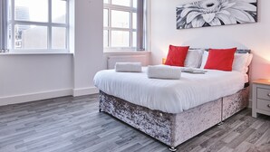 A bedroom with a large bed, white bedding, a red and white pillow set, a gray and white patterned headboard, a nightstand with a lamp, and a large window with a view of buildings. - Winter Gardens Service Apartments 2 (Blackpool)