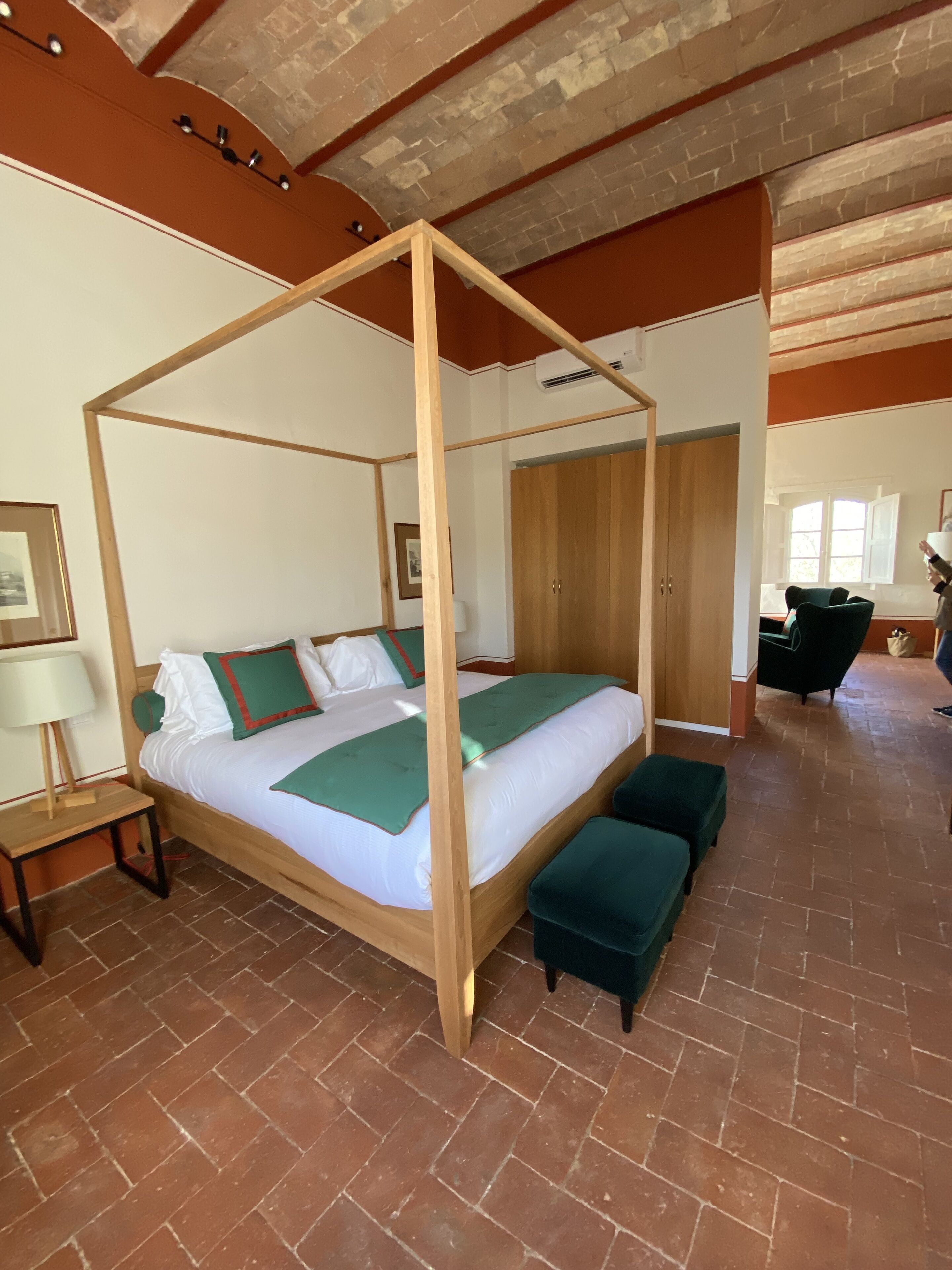 Een hemelbed met houten frame, een groene plaid en bijpassende kussens. Er is een klein nachtkastje met een lamp, een bankje aan het voeteneinde van het bed, en een kastje op de achtergrond.