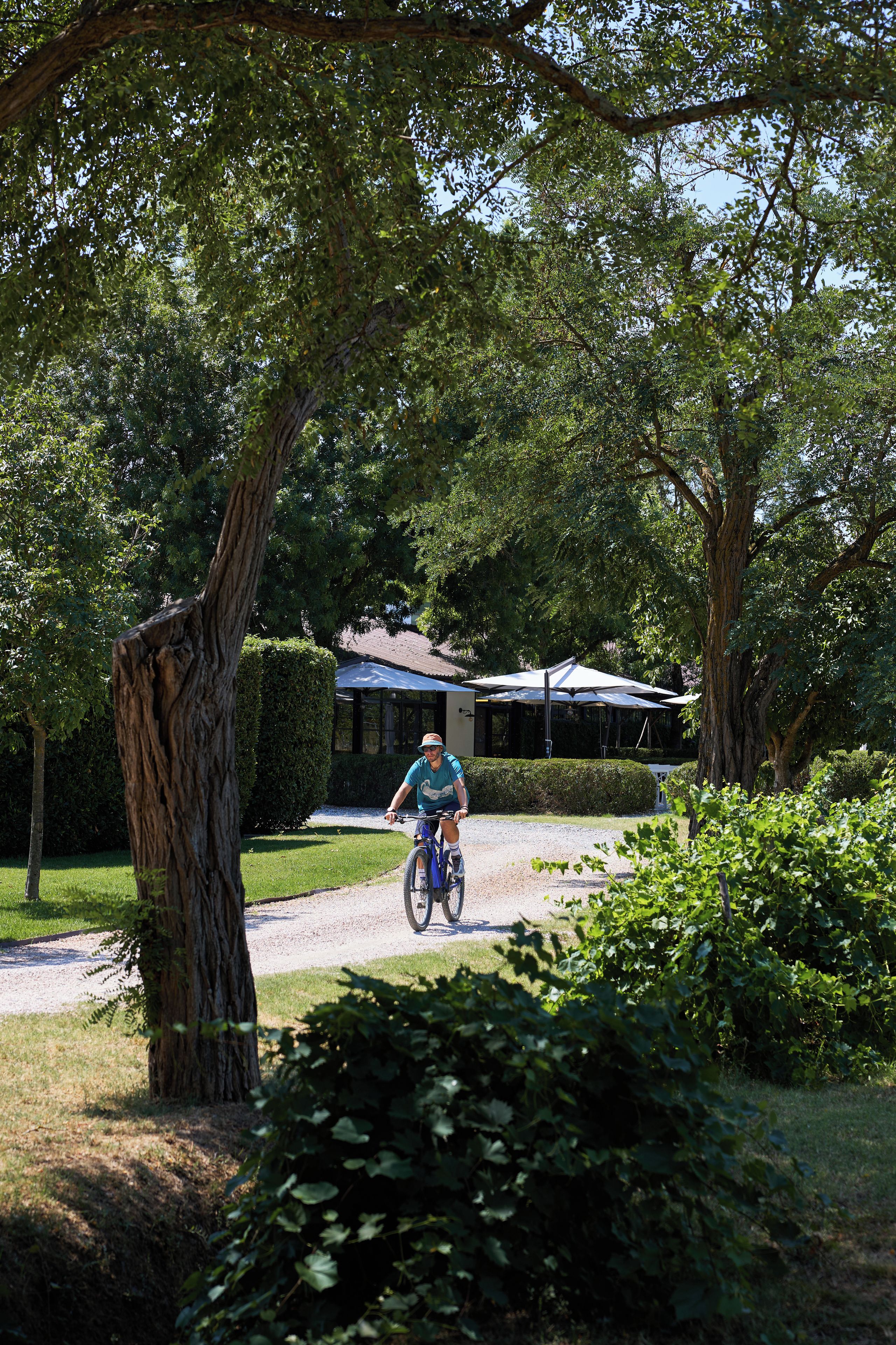 A person riding a bicycle on a gravel path in a lush, green area with trees and a building in the background.