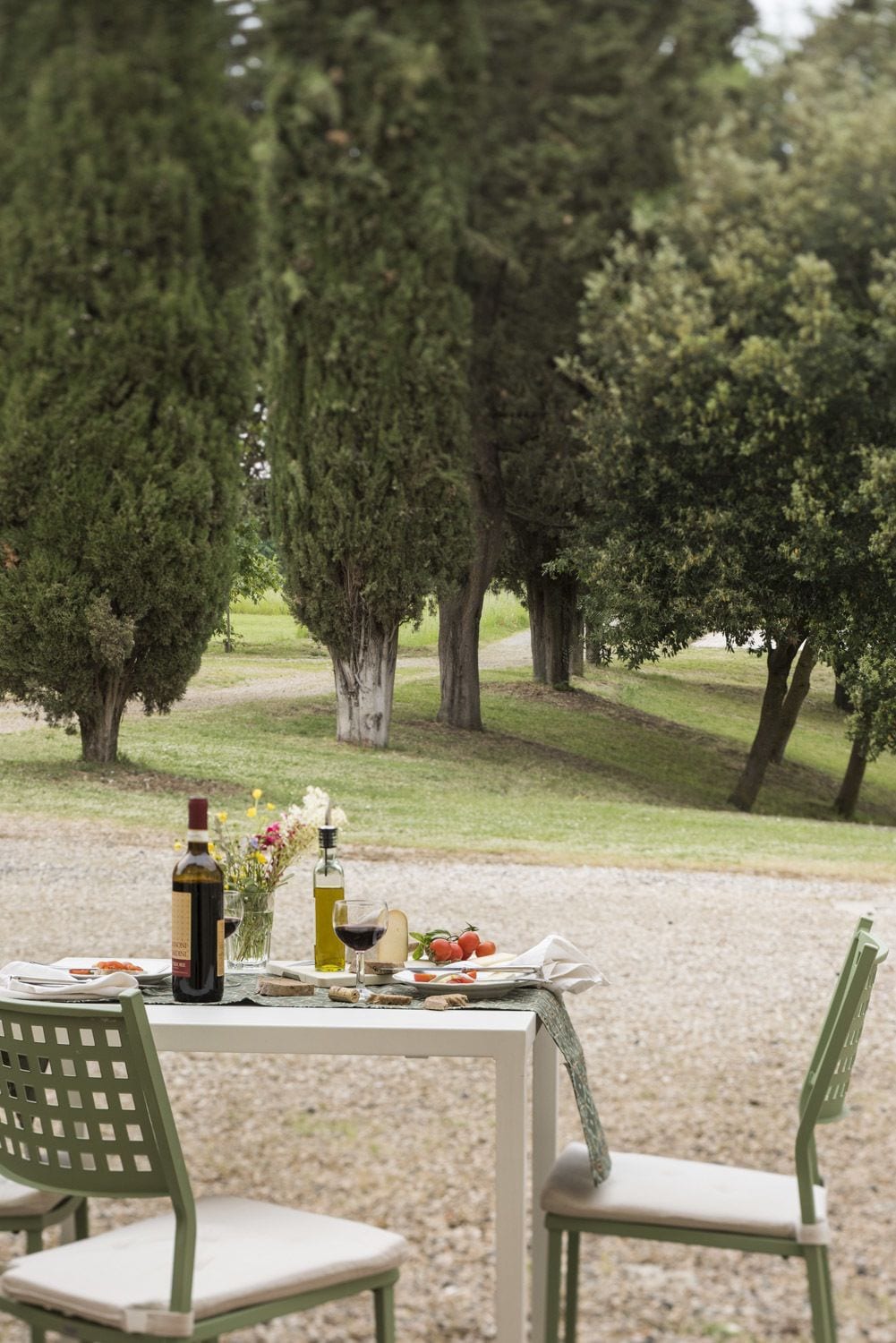 An outdoor dining table with a bottle of wine, a glass of wine, a bottle of olive oil, a plate of food, and a vase of flowers.