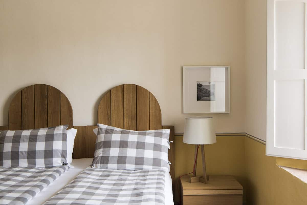 Hotelkamer met een bed, twee geruit kussens, een houten hoofdbord, een nachtkastje met een lamp, en een ingelijst schilderij aan de muur.