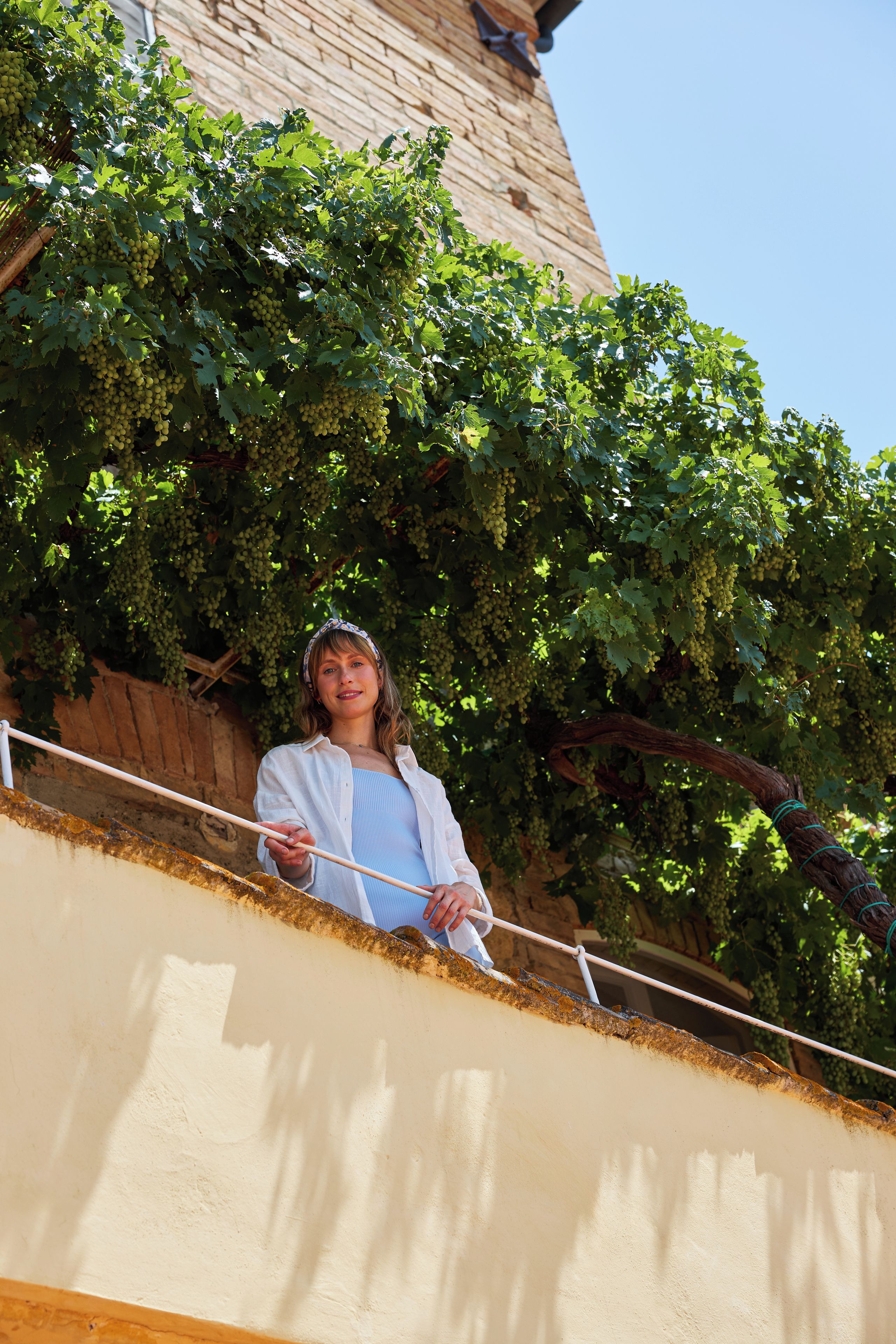 Eine Person steht auf einem Balkon mit Blick auf einen Weinberg und ein Backsteinhaus im Hintergrund.