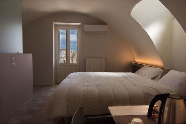 A bedroom with a bed, a table with a kettle, and a window with a view of buildings.
