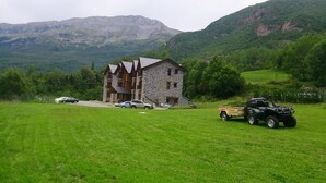 Un edificio de piedra con techo inclinado, rodeado de un campo cubierto de hierba y montañas al fondo.