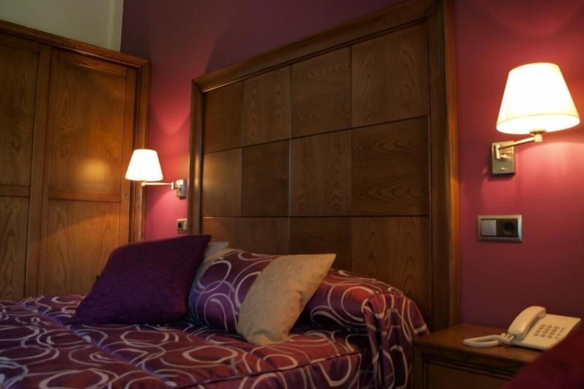 A hotel room with a wooden headboard, a bed with patterned bedding, two wall-mounted lamps, and a bedside telephone.