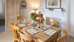 A dining room with a wooden table set for six, surrounded by wooden chairs. A vase with tulips is on the table, and a mirror hangs on the wall above it.