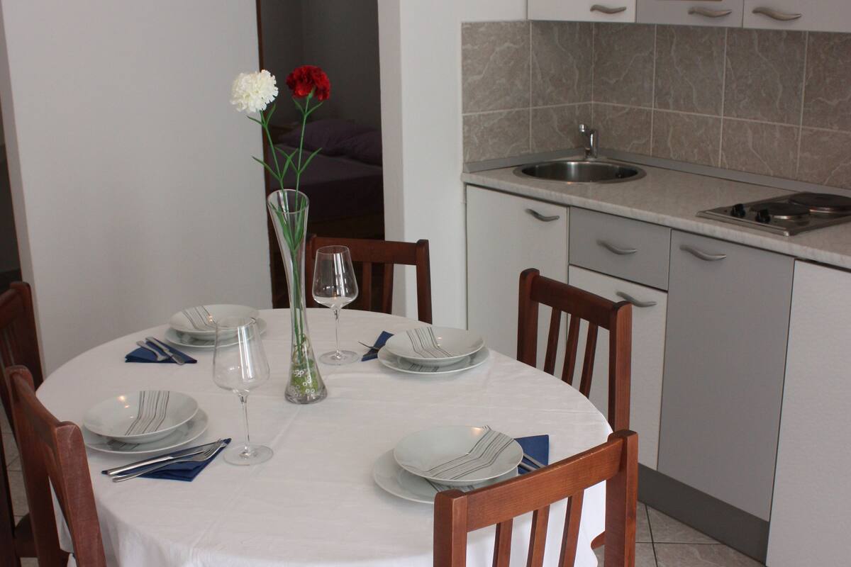 A dining area with a round table set for four, surrounded by wooden chairs. A vase with flowers is placed on the table.