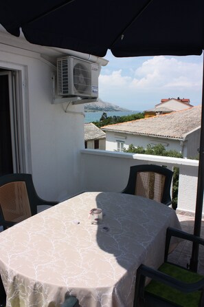 Un balcon extérieur avec une table et des chaises, un appareil de climatisation et une vue sur des bâtiments et une étendue d’eau en arrière-plan.
