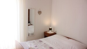 Ein ordentlich bezogenes Bett mit weißer Decke und einem gerollten Handtuch, ein Nachttisch mit Lampe, ein Spiegel an der Wand und eine herzförmige Dekoration.