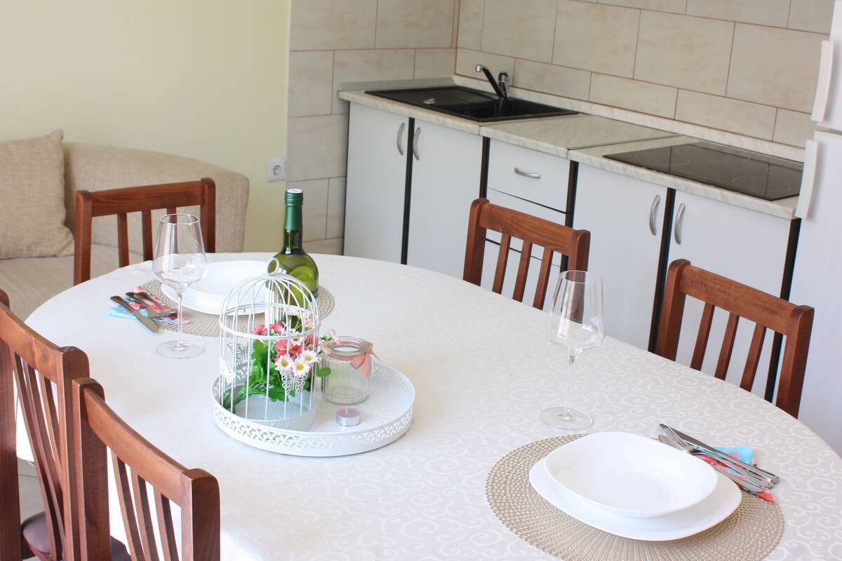 A dining area with a round table set for four, surrounded by wooden chairs. A white tablecloth covers the table, and there are plates, cutlery, and a bottle of wine on it. The room has white cabinets and a tiled backsplash.