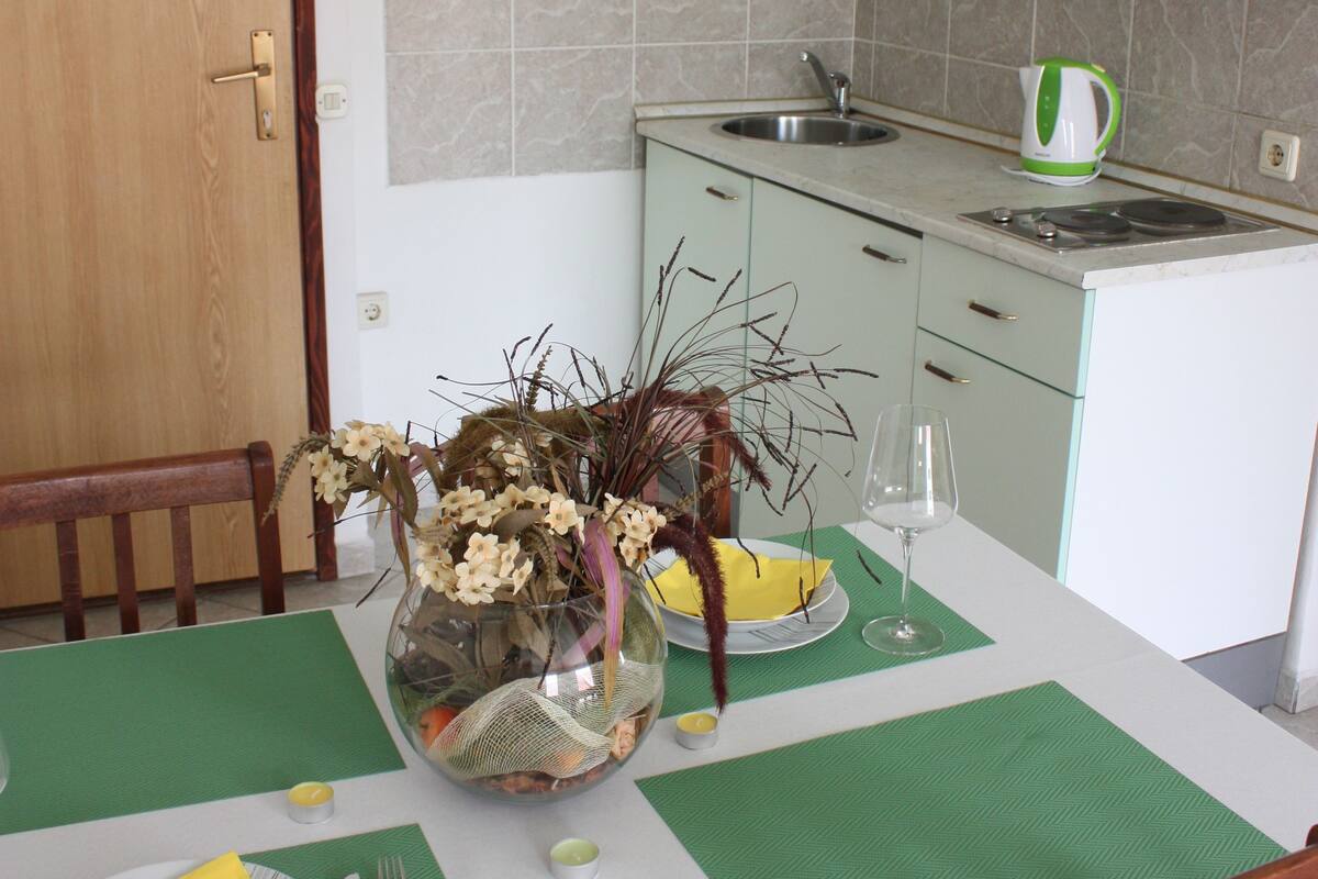 A dining area with a table set for two, a vase with flowers, and a kitchen in the background.