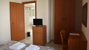 Un dormitorio con una cama, un armario de madera, un escritorio con silla, un televisor sobre un mueble bajo y una puerta que da al baño.