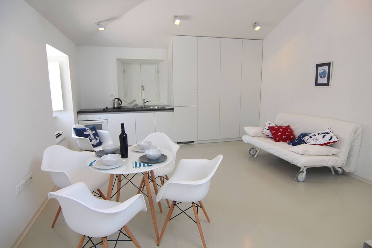 Cozinha moderna com armários brancos, uma mesa de jantar com cadeiras e um sofá branco com almofadas coloridas.