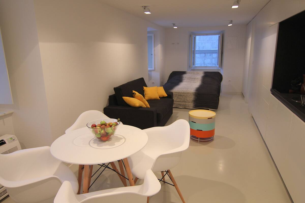 Uma sala de estar moderna com uma mesa branca, um sofá preto com almofadas amarelas e uma mesinha lateral colorida.
