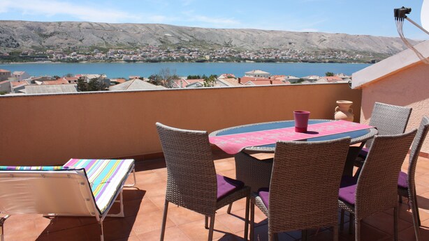 Een terras met rieten stoelen en een tafel, met uitzicht op een meer en stad.