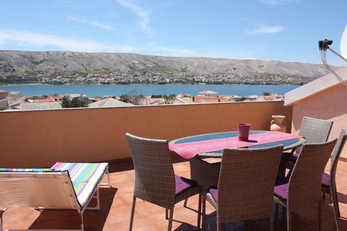 A terrace with wicker chairs and a table, overlooking a lake and town.