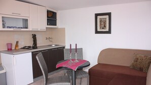 Een gezellig compact verblijf met een kitchenette, een eethoek met twee stoelen, een bank en een foto in een lijst aan de muur.