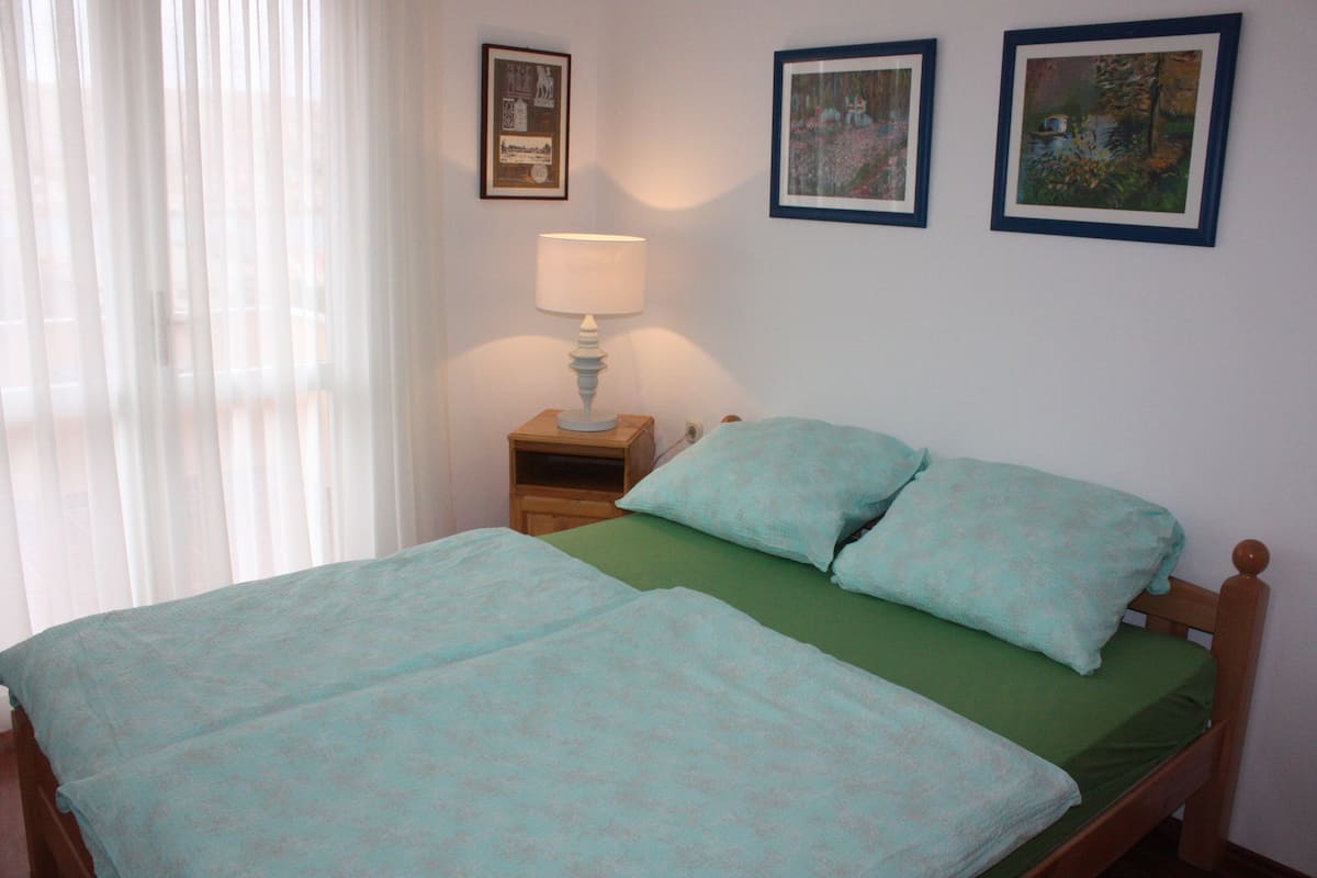 A double bed with green and light blue bedding, a bedside table with a lamp, and two framed pictures on the wall.