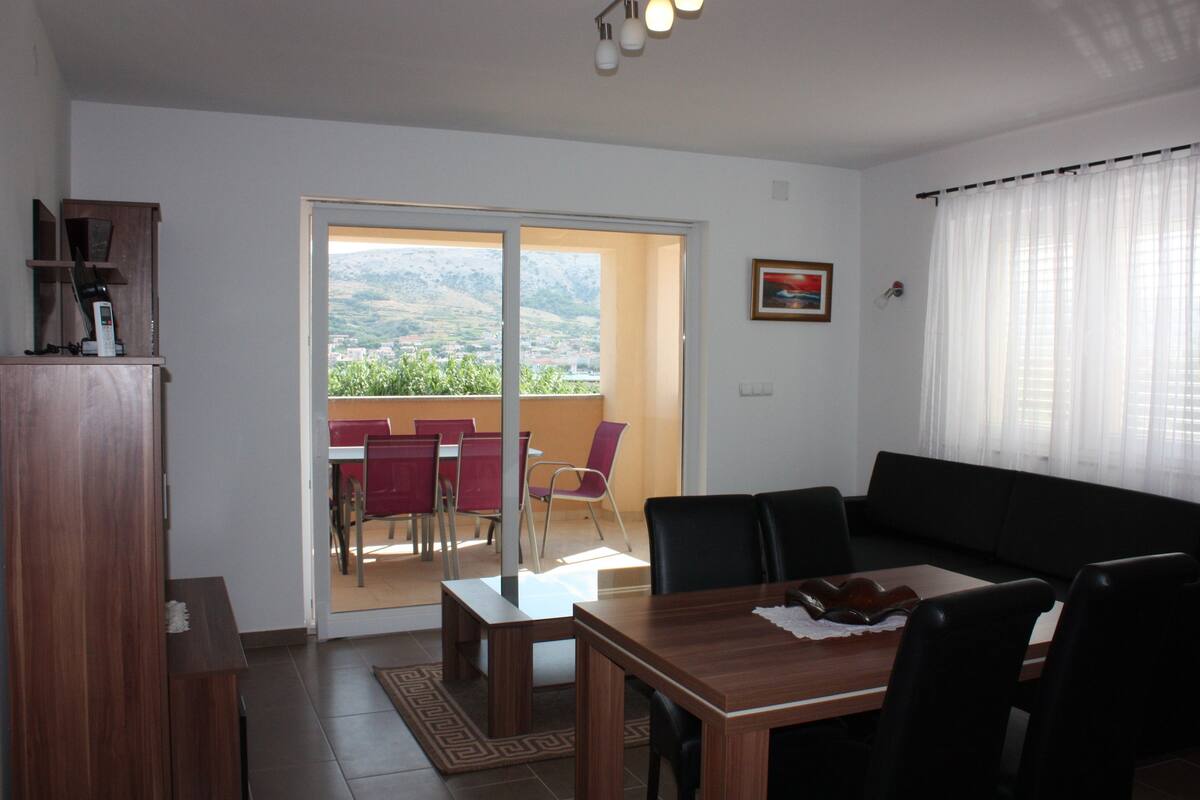 A living room with a sofa, a dining table, and a balcony with a view of the mountains.