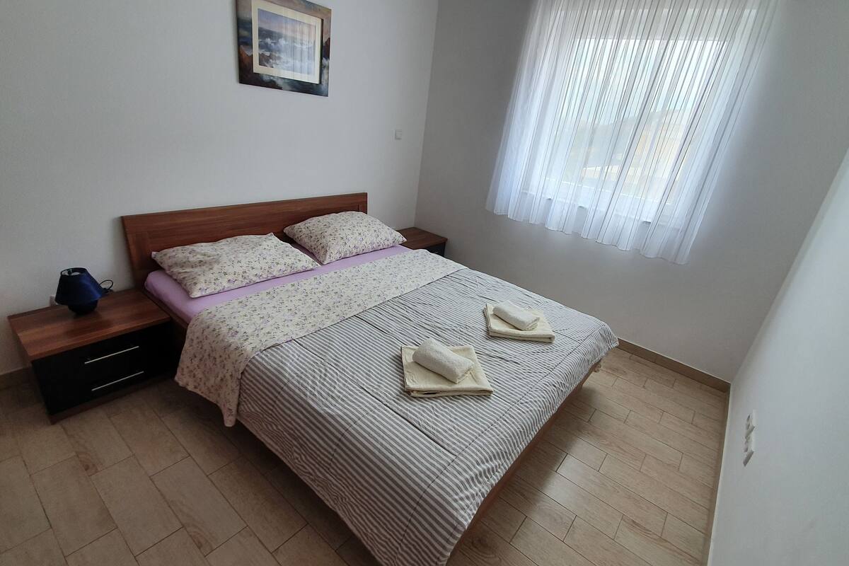 A neatly made bed with a striped bedspread and two pillows, a bedside table with a lamp, and a window with sheer curtains.
