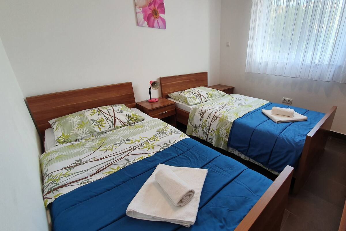 A room with two beds, each with a blue bedspread and a bamboo-themed pillow. There is a nightstand with a lamp and a power outlet on the wall.
