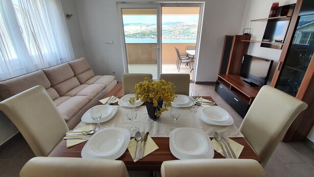 Uma área de jantar com uma mesa posta para quatro pessoas, um sofá, uma televisão e uma varanda com vista.