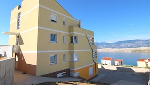 A multi-story building with a sloped roof, a balcony, and a view of a body of water and mountains in the background. - Apartments Ljubic (Pag)