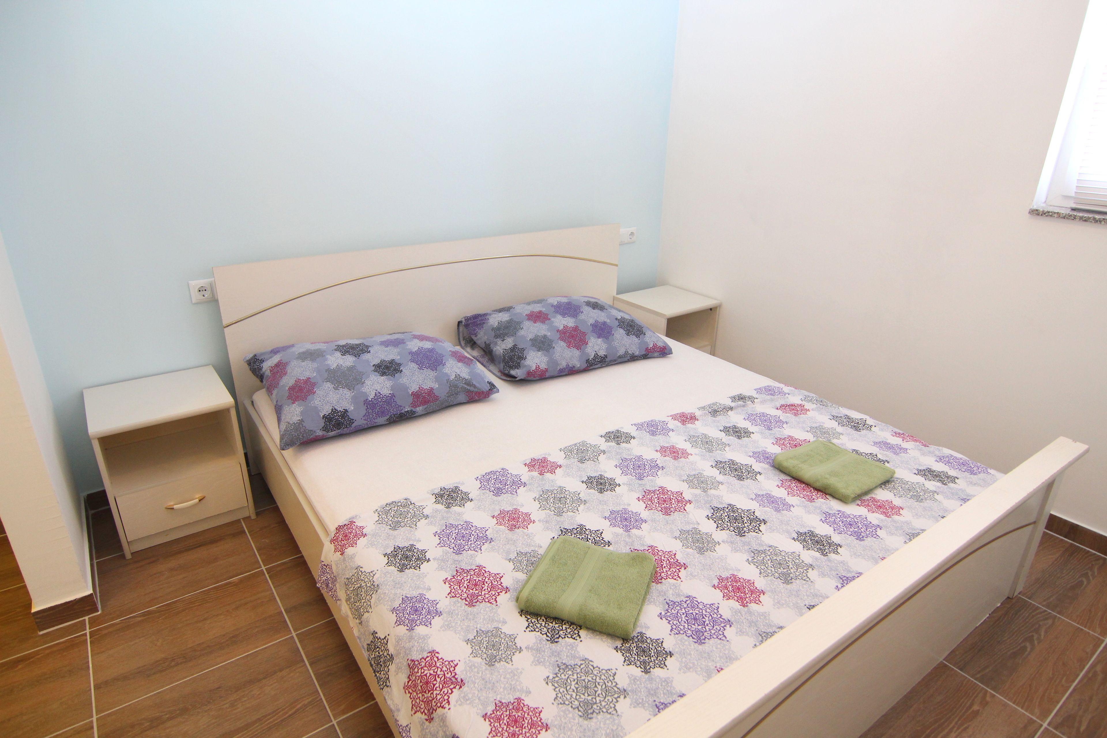 A neatly made bed with patterned bedding, two pillows, a green towel, and a bedside table with a drawer.