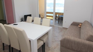 A dining area with a white table and chairs, a sofa, and a view of the sea through a glass door. - Apartments Ljubic (Pag)