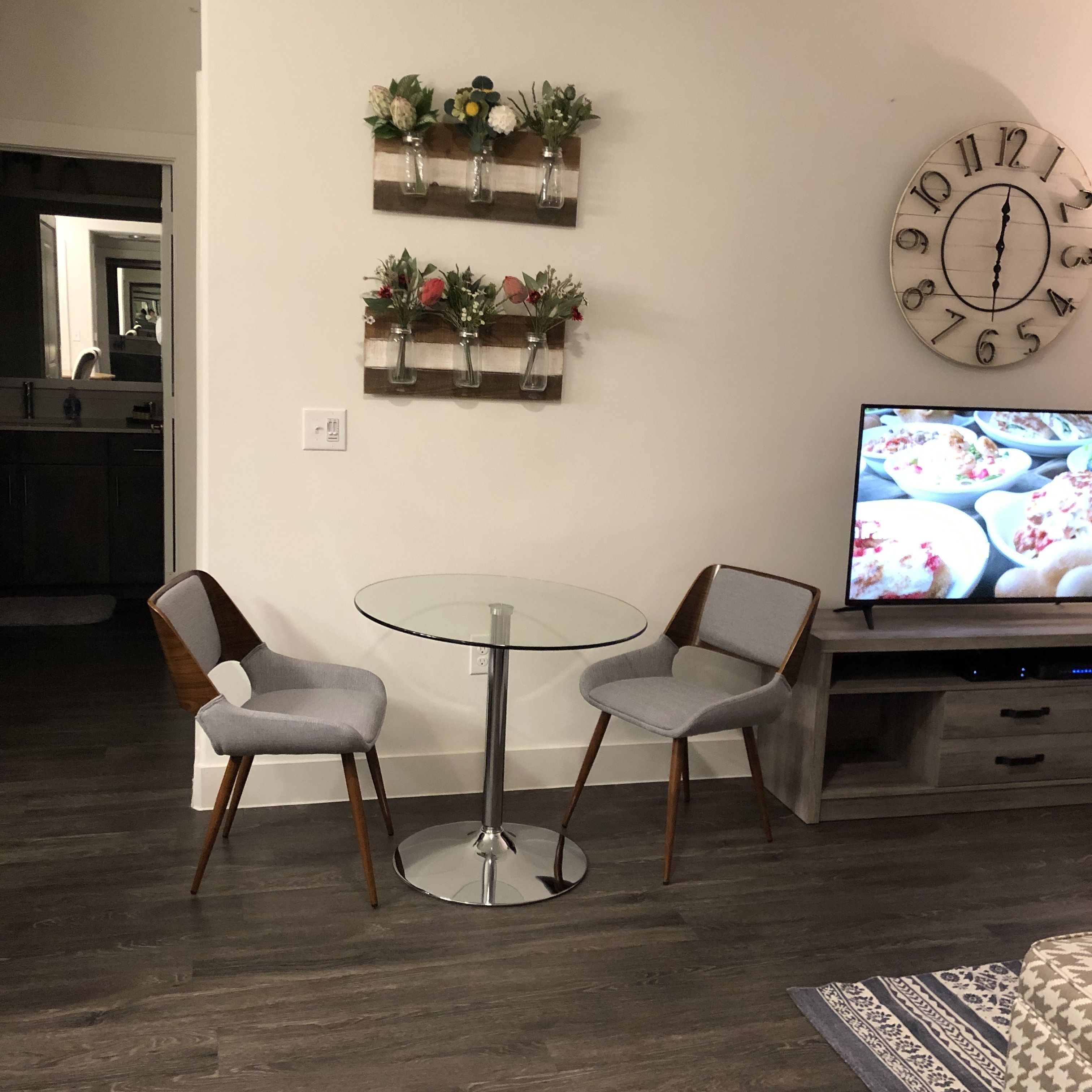 A dining area with a glass table and two chairs, a wall clock, and a television displaying food.