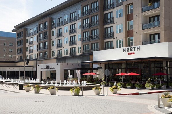 Un hôtel à plusieurs étages, doté d’une façade moderne, d’un espace extérieur aménagé avec des parasols rouges et d’un élément décoratif aquatique à l’avant.