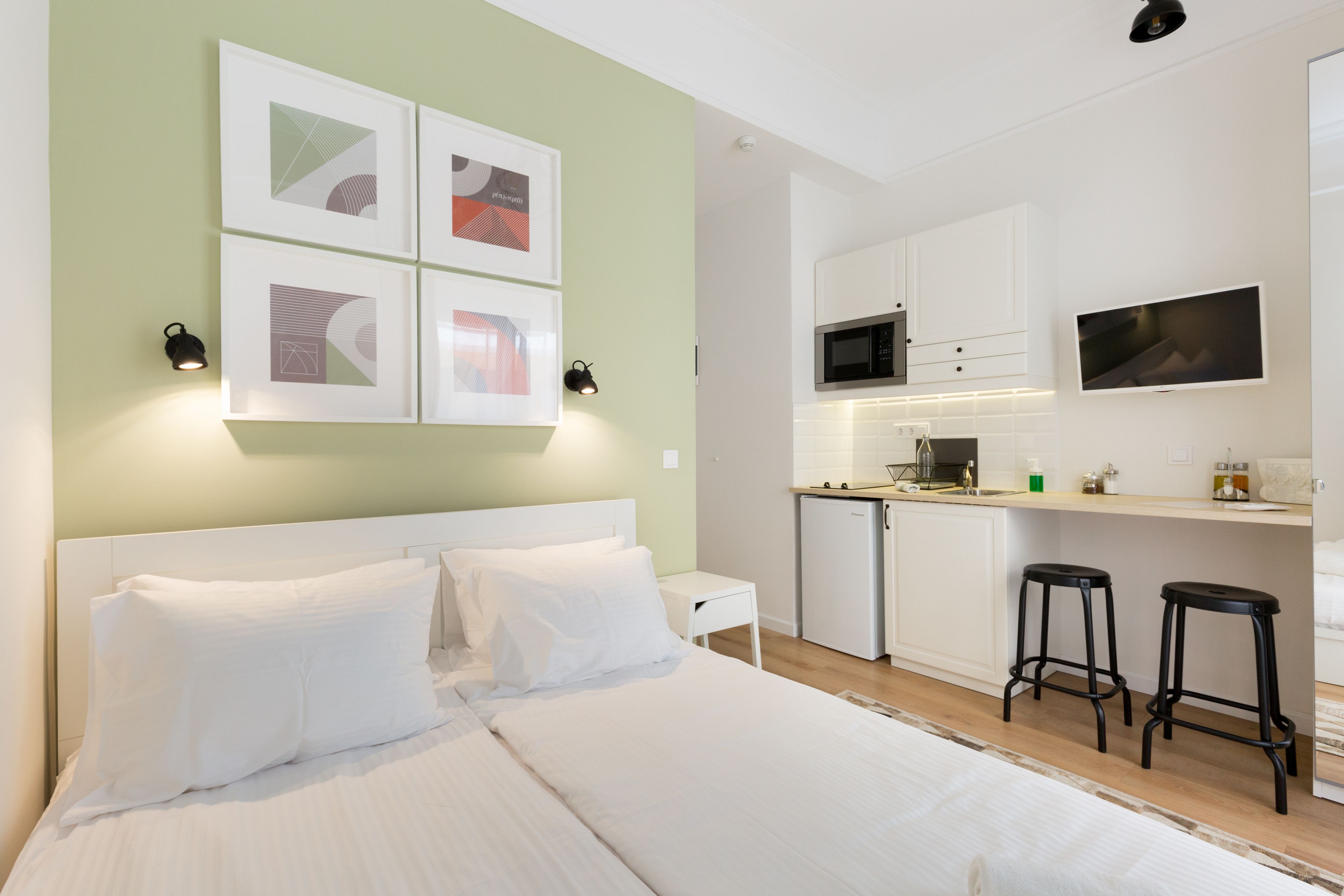 A modern hotel room with a large bed, a kitchenette, and a TV mounted on the wall.