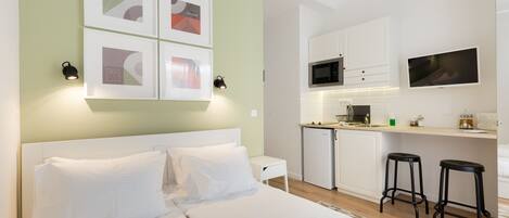 A modern hotel room with a large bed, a kitchenette, and a TV mounted on the wall.