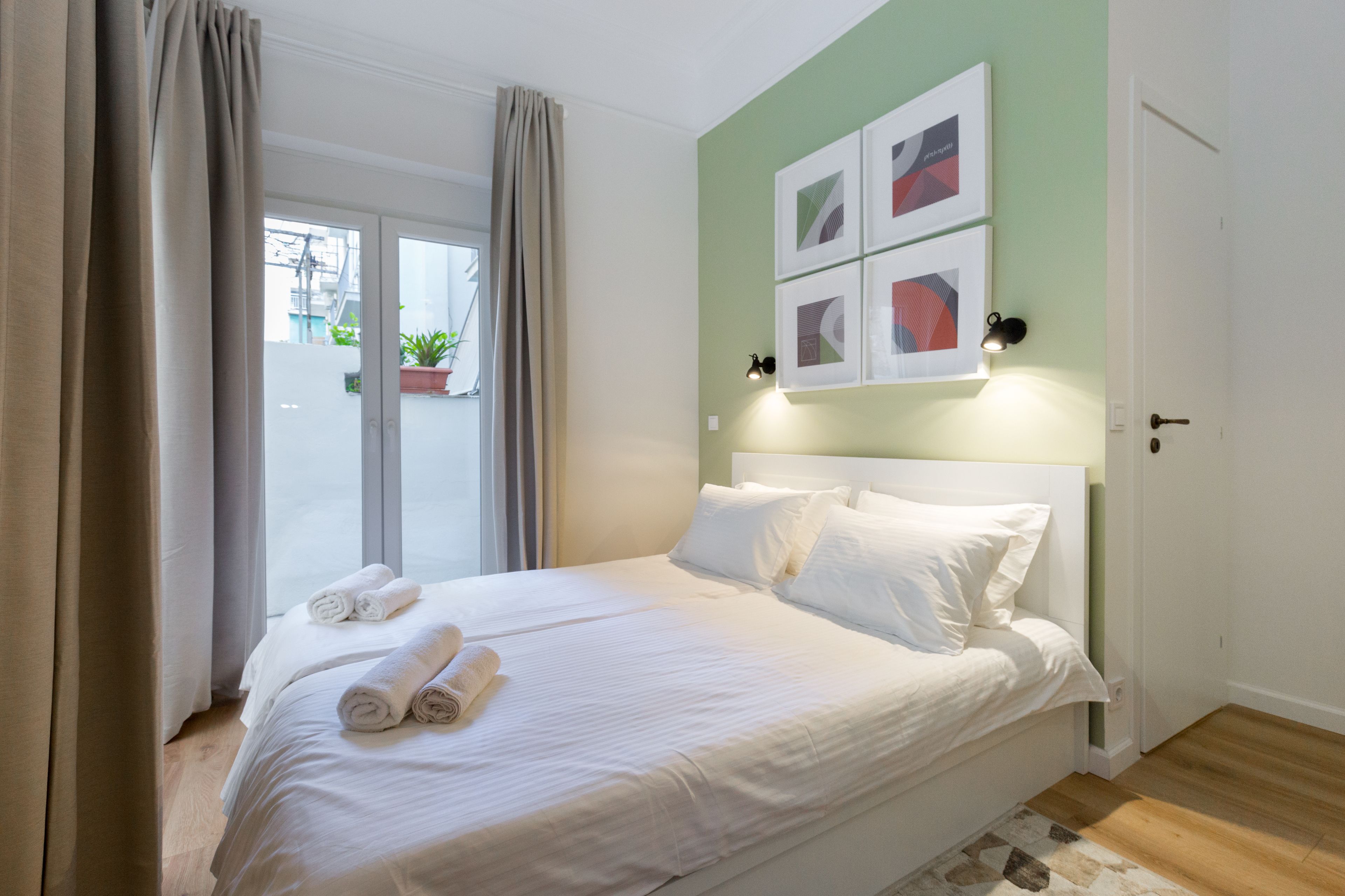 A neatly made bed with white linens and rolled towels, a green accent wall, and a view through a glass door.