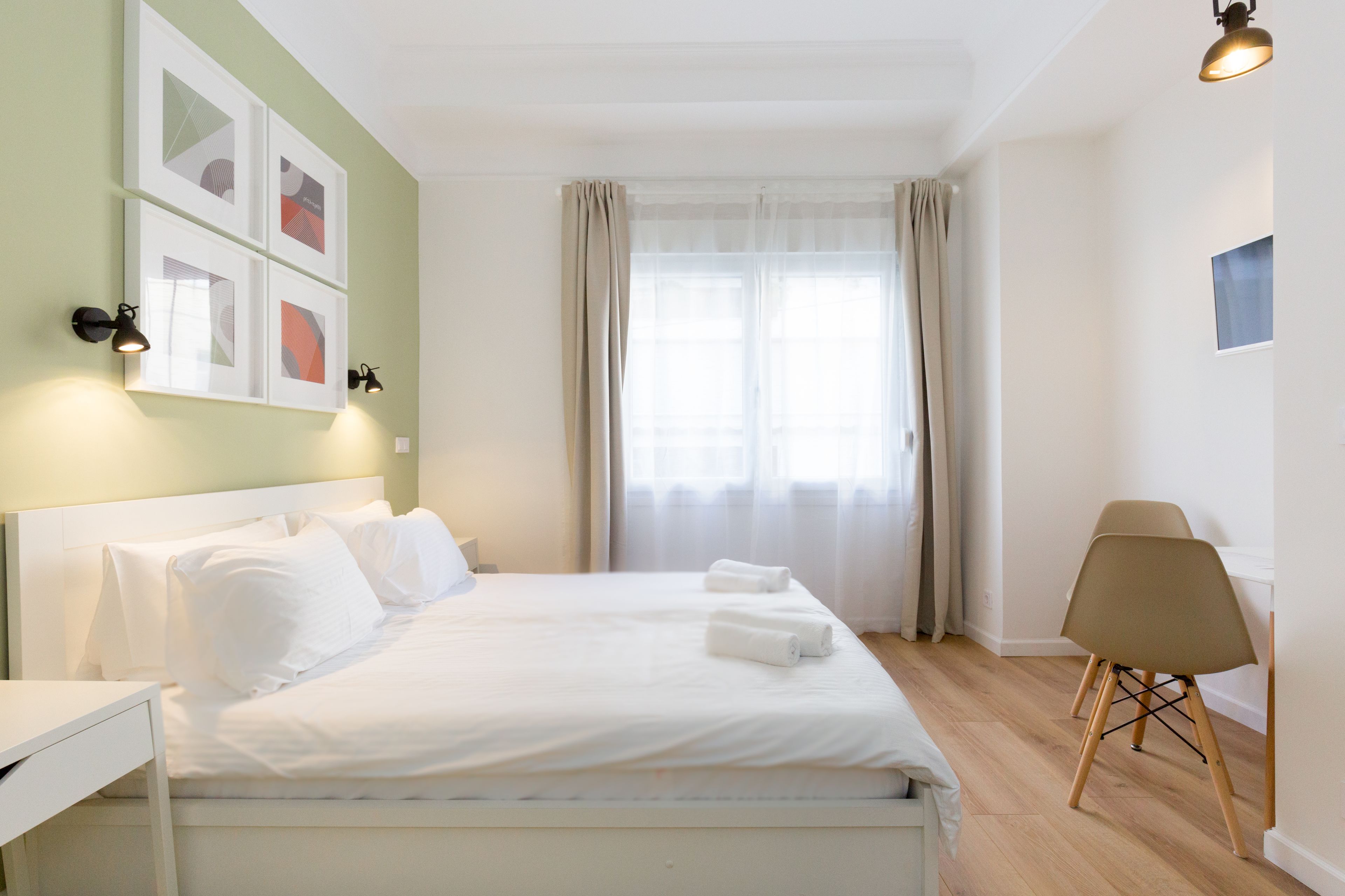 A neatly made bed with white linens and two rolled towels, a bedside table, a chair, and a window with sheer curtains.